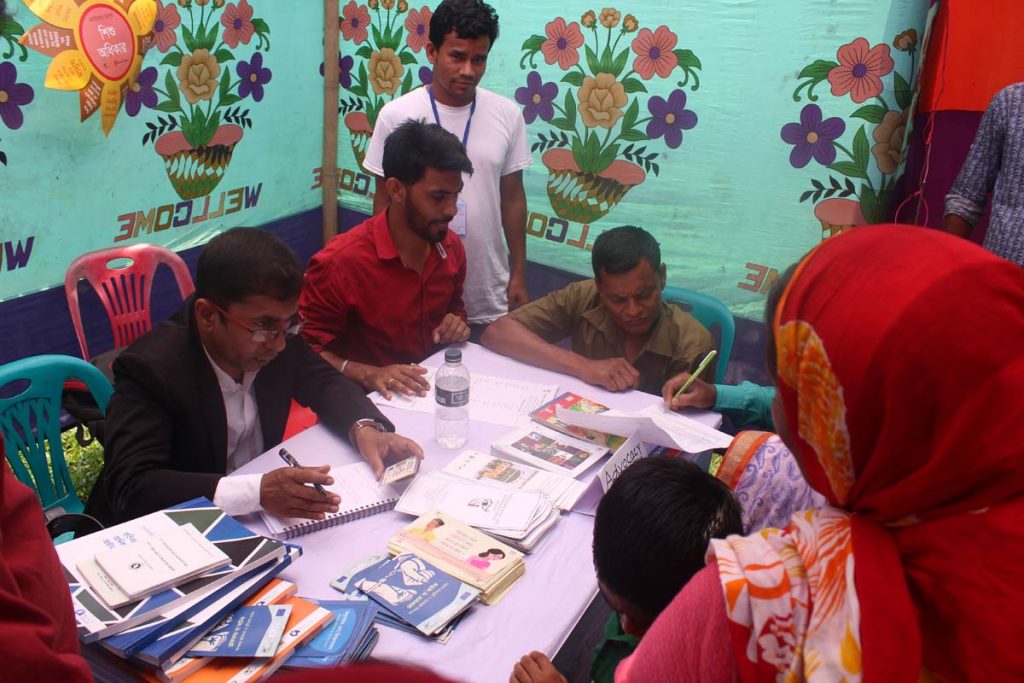 A lawyer dressed in a traditional black blazer and white collared shirt sits at a table covered with educational materials in a brightly colored tent and listens to a woman seated across the table.